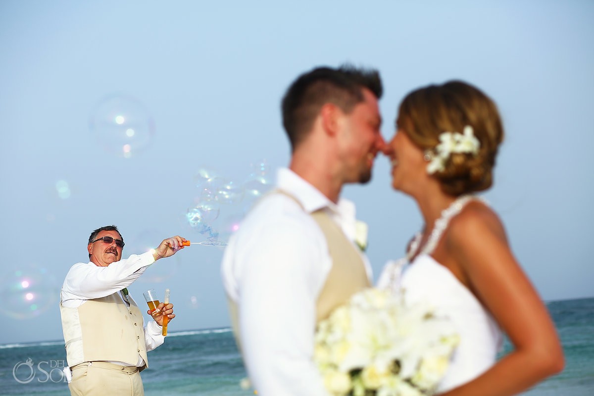 father blows bubbles in the background as bride and groom kiss at dreams tulum wedding