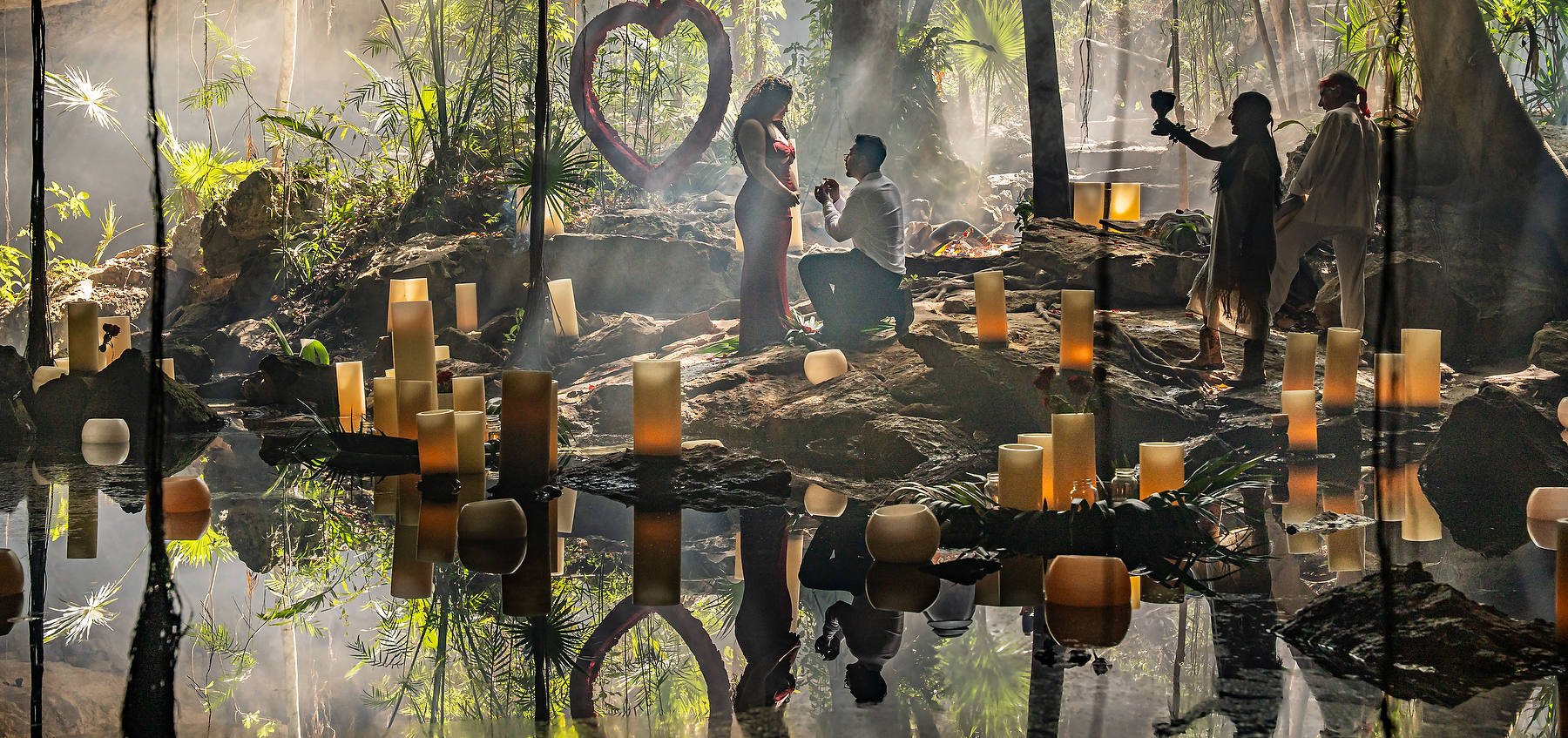 This is an example of a creative proposal idea for men, Groom proposing on his knee to ask for her hand, she said yes, then they had a wedding and married in the same moment, the cenote decoration includes candles and a floating heart made of red roses, mayan ceremony offering a copal blessing with a spiritual mayan wedding ceremony, reflections of cenote candle decoration reflecting in the waters of the underground river. Photo by Matt Adcock