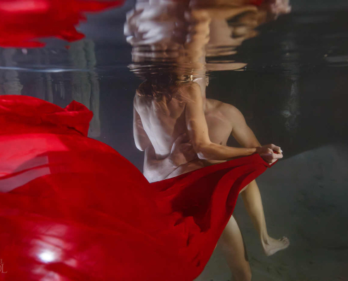Artistic underwater cenote couple session featuring flowing red fabric and intimate connection inside a cave inspired by Adam and Eve