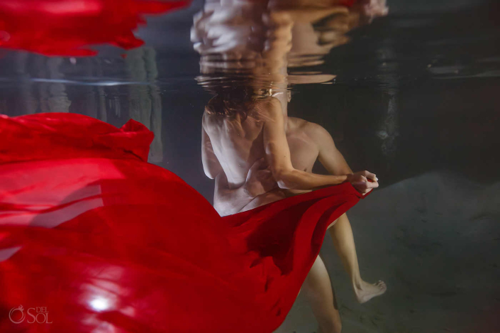 Underwater Trash the Dress Cenote Session Artistic underwater cenote couple session featuring flowing red fabric and intimate connection inside a cave inspired by Adam and Eve