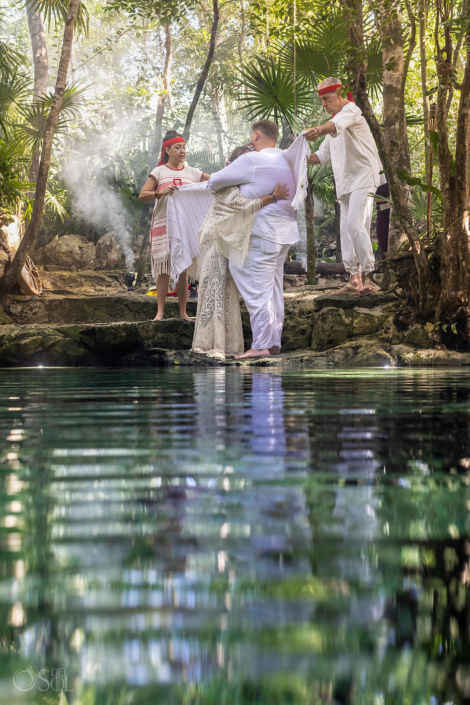 Couple wrapped together in a ceremonial shawl beside the water during their cenote wedding